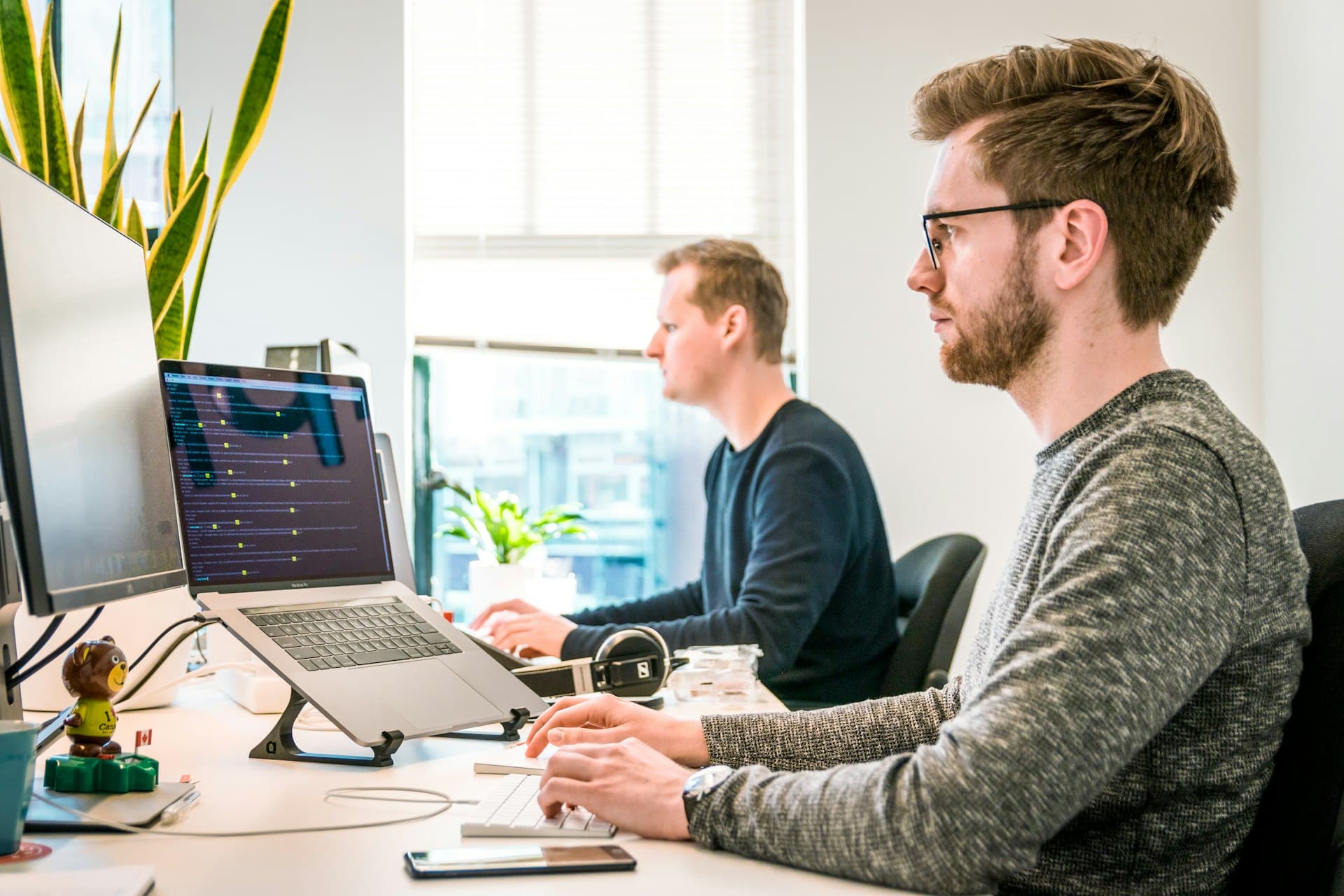 Desarrollador programando en su laptop en una oficina, con otro programador al fondo